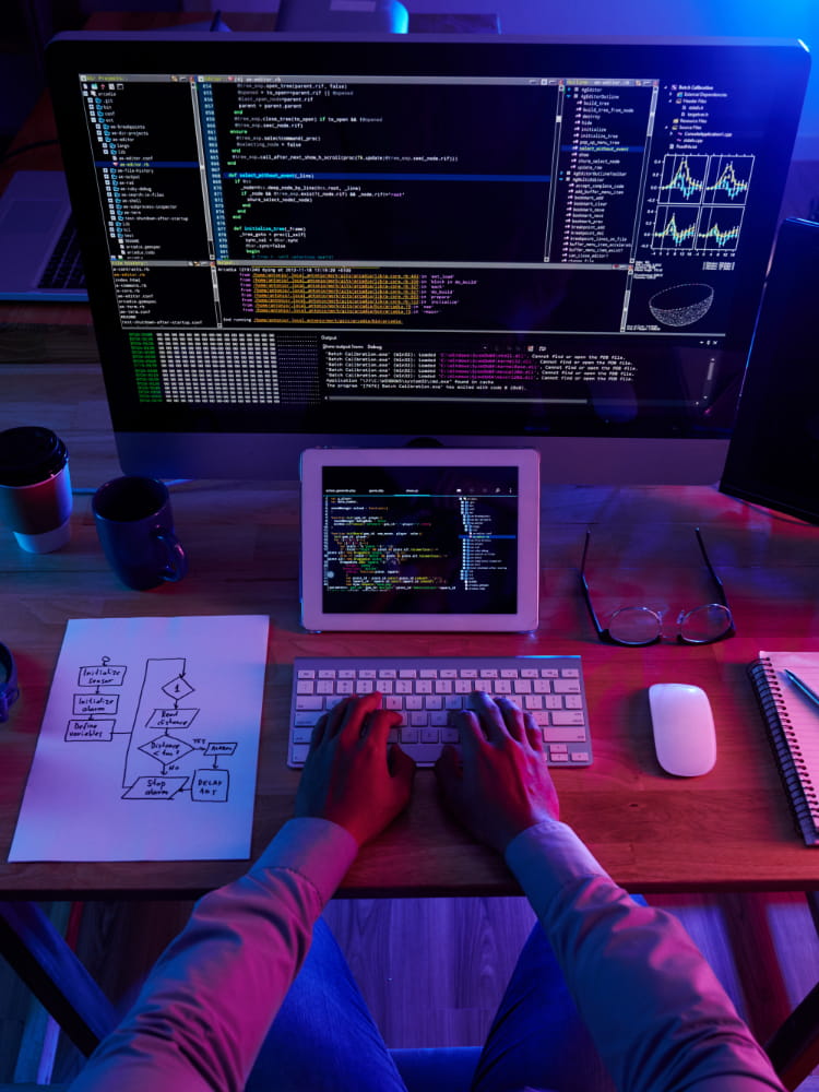A modern programming workspace featuring a large monitor displaying multiple code windows, terminal outputs, and graphical plots. A tablet beside it shows additional code. On the wooden desk are a printed flowchart diagram, glasses, a mouse, a notebook with a pen, and two coffee mugs. The scene is lit with blue and pink tones, creating a focused, tech‑driven atmosphere.