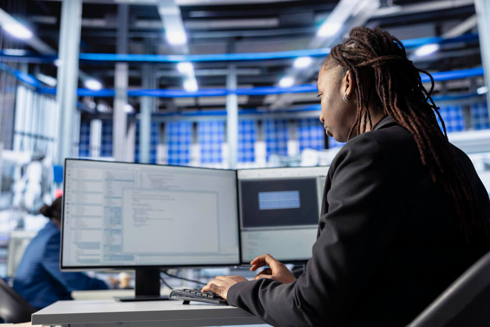 A person working at a desk in a modern, high-tech office, focused on two large monitors displaying code and data visualizations. The workspace features an industrial-style ceiling with blue accent lighting and multiple workstations, suggesting a collaborative, technologically advanced environment.