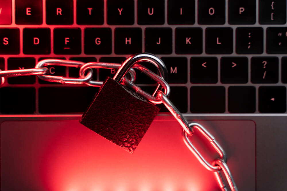 Close-up of a laptop keyboard wrapped in a heavy metal chain with a padlock, illuminated by red lighting. The dramatic setup symbolizes cybersecurity, data protection, and restricted access to digital information.