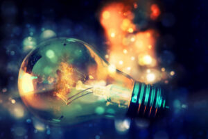 A glowing light bulb with a visible filament, set against a vibrant abstract background featuring bokeh effects in blue, orange, and yellow hues—symbolising creativity, energy, and illumination.