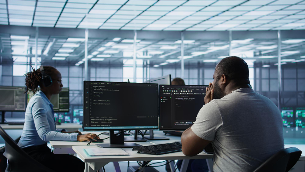 Two people working at computer stations in a modern tech office, with multiple monitors displaying programming code and development tools.