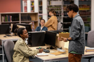 Students working in a library or study area, with one seated at a desk using a calculator and notebook, while another stands nearby holding a notebook and engaging in discussion. In the background, two others are visible near bookshelves and computer desks, contributing to a focused academic atmosphere.
