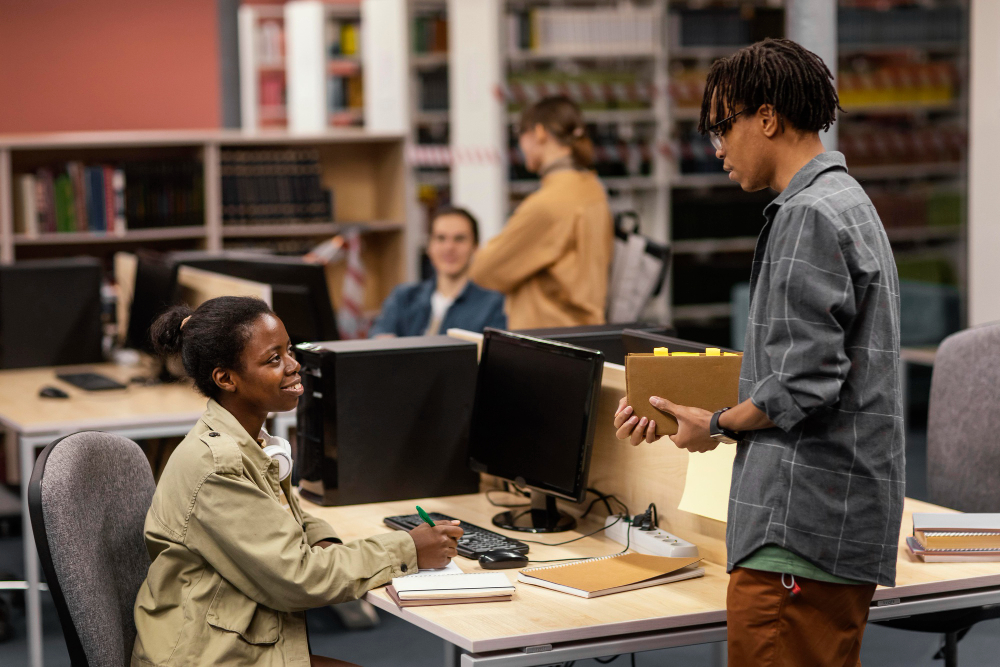 Students working in a library or study area, with one seated at a desk using a calculator and notebook, while another stands nearby holding a notebook and engaging in discussion. In the background, two others are visible near bookshelves and computer desks, contributing to a focused academic atmosphere.