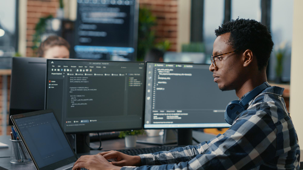 Person working at a desk with multiple monitors displaying programming code and a terminal interface, suggesting software engineering in a modern office setting.