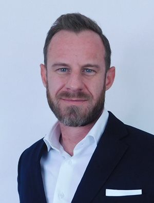 Man in a dark suit jacket and white dress shirt, with short hair and a beard, standing against a plain white background, looking directly at the camera with a neutral expression.