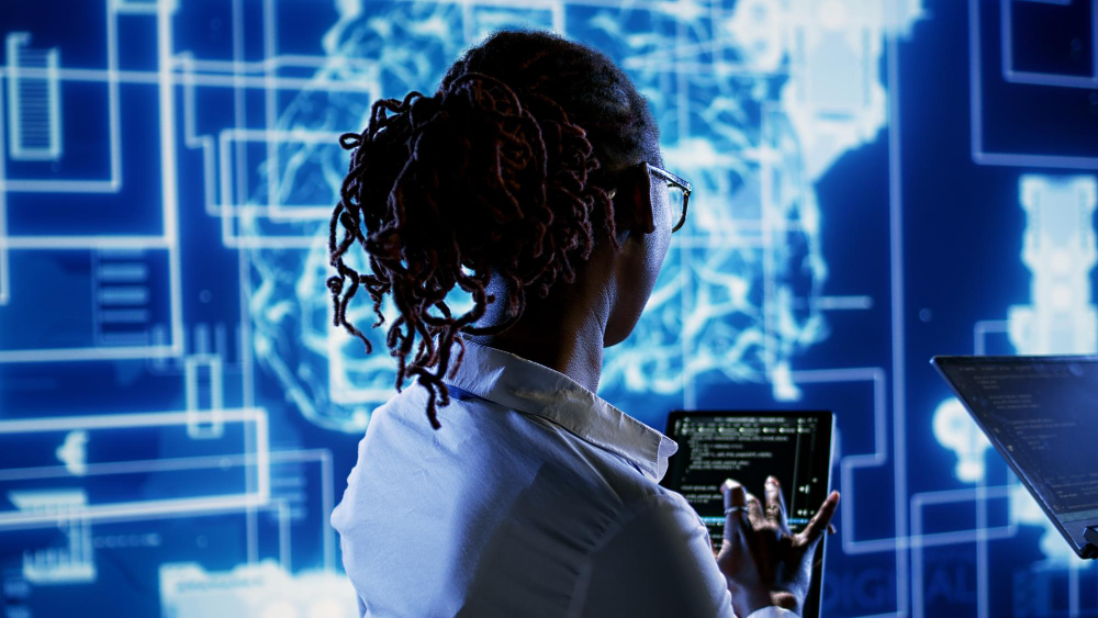 A person with dreadlocks and glasses, wearing a white shirt, stands in a high-tech workspace. They hold a tablet displaying code while facing a large digital screen featuring a stylized brain surrounded by data visualizations and interface elements. A nearby laptop also shows lines of code, suggesting a focus on artificial intelligence or machine learning.