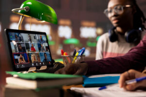 A tablet on a cluttered desk displays a virtual meeting with multiple participants, while two people in the foreground engage with the screen and take notes, suggesting collaborative online learning in a study environment.