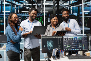 Four professionals stand in a modern server room, discussing data on a laptop and tablet. Computer monitors display cybersecurity visuals and network diagrams, while rows of servers with blinking lights form the high-tech backdrop. The scene highlights teamwork and digital infrastructure management in an IT environment.
