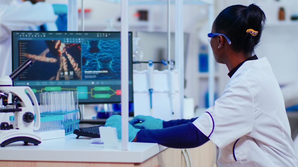 A scientist in a white lab coat, gloves, and goggles works at a computer in a modern laboratory. The screen displays a close-up of a DNA strand labeled "Double-stranded DNA" and a colorful flowchart. Nearby, test tubes are organized in racks, and a microscope sits on the bench, suggesting genetic or molecular biology research.