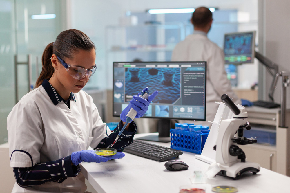 Two researchers work in a clean, modern laboratory. In the foreground, one uses a pipette to transfer liquid into a petri dish while seated at a workstation with a microscope, test tubes, and a computer displaying microscopic images. In the background, another scientist works at a similar setup, highlighting collaborative scientific research and advanced lab technology