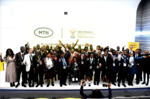 A large group of students in school uniforms and adults in formal attire pose together in front of a branded backdrop featuring the MTN and Department of Basic Education logos. The backdrop reads “Ministerial Breakfast with the Matric Class of 2022,” indicating a celebratory event recognizing South African matriculants. The mood is formal and festive, highlighting partnership and achievement.