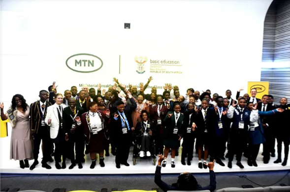 A large group of students in school uniforms and adults in formal attire pose together in front of a branded backdrop featuring the MTN and Department of Basic Education logos. The backdrop reads “Ministerial Breakfast with the Matric Class of 2022,” indicating a celebratory event recognizing South African matriculants. The mood is formal and festive, highlighting partnership and achievement.