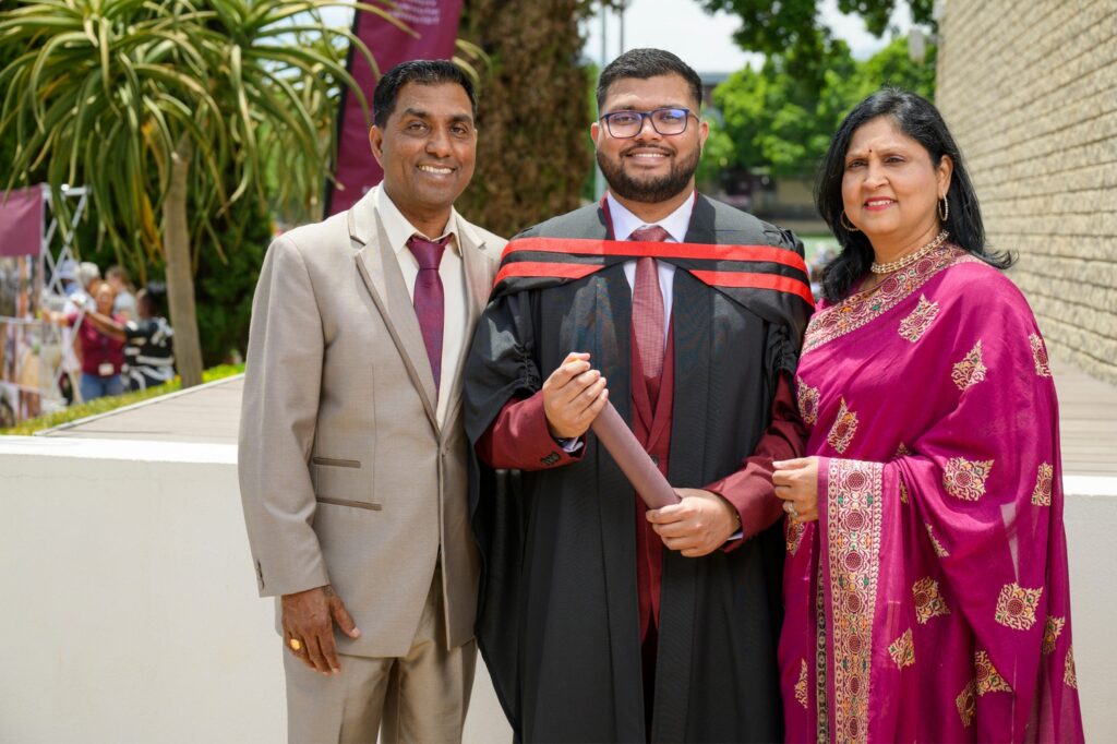 A graduate in black academic regalia with red trim stands proudly holding a diploma, flanked by two formally dressed family members — one in a beige suit and maroon tie, the other in a maroon and gold traditional outfit. The outdoor setting includes greenery, a stone wall, and other people in the background, suggesting a celebratory university graduation event.