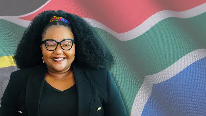A smiling person with voluminous curly hair and large black-framed glasses stands confidently in front of a vibrant South African flag. They wear a black blazer with gold buttons over a black top, conveying a polished and professional look. The flag’s bold colors and Y-shape design emphasize national pride and cultural identity.