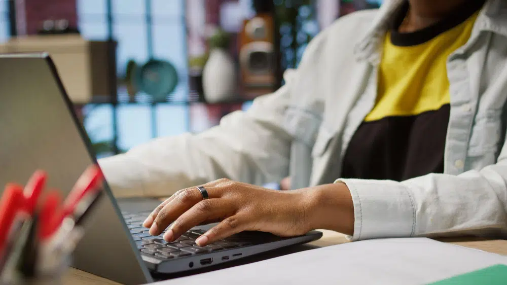 Alt text: A person sits at a desk typing on a laptop, wearing a light shirt over a black-and-yellow top. Their hands rest on the keyboard, one with a ring. On the desk are red pens in a holder and scattered papers. Behind them is a window with a grid pattern and shelves holding plates and small plants, creating a modern, home‑office feel