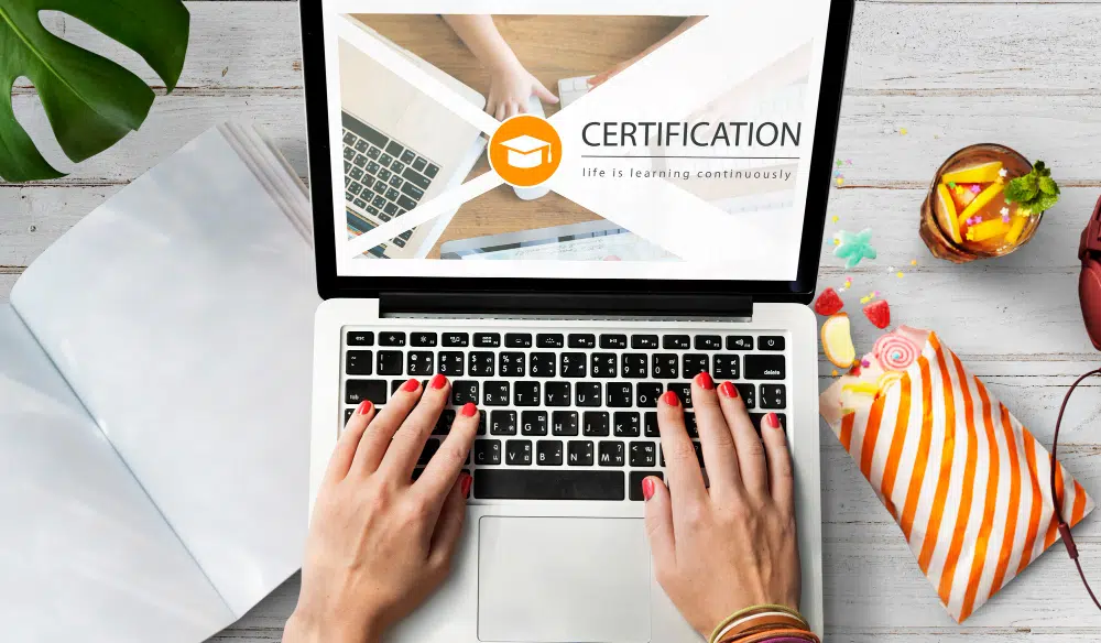 A person types on a laptop displaying a certification graphic with a graduation‑cap icon and the words “CERTIFICATION – life is learning continuously.” The laptop sits on a white wooden surface surrounded by an open notebook, a tropical drink with fruit garnishes, a striped bag, and a green plant, creating a relaxed, summery study environment.