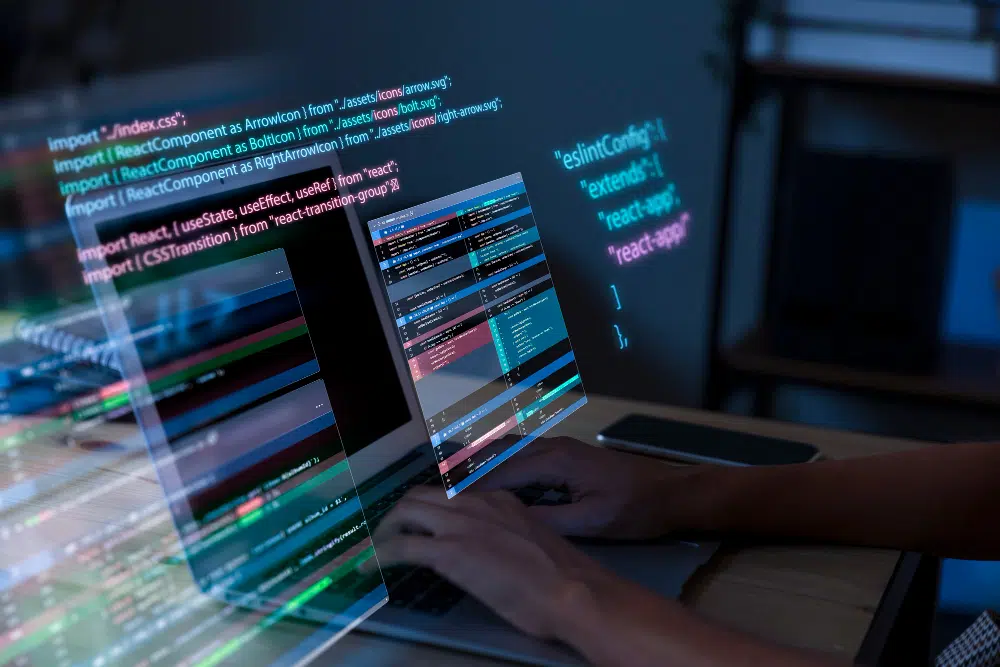 A person works on a laptop displaying multiple screens of code, including React.js components and configuration files. The setup features syntax‑highlighted JavaScript, import statements, and terminal output, creating a focused software‑development workspace.