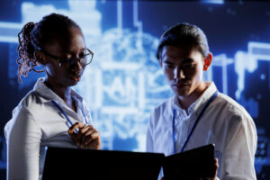Two people wearing white shirts and lanyards stand together, examining their devices while a large blue digital schematic glows on the screen behind them, creating a focused, high‑tech workspace.