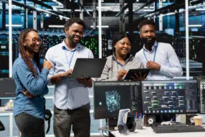 A group of four IT professionals stand together in a modern server room, discussing work while holding a laptop and a tablet. Multiple monitors in the foreground display network diagrams and data visualisations, and illuminated server racks fill the background, highlighting a high‑tech, collaborative environment.