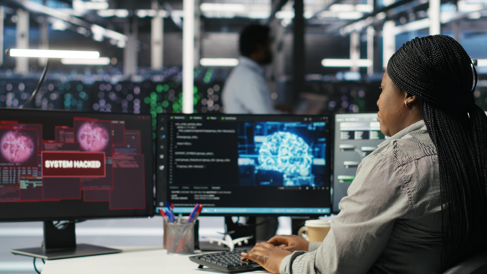 A cybersecurity professional sits at a desk monitoring multiple screens, including one displaying a red “SYSTEM HACKED” alert over digital maps and data. Another screen shows code and a digital brain graphic, suggesting AI‑driven analysis. A server room with illuminated equipment is visible in the background, highlighting a high‑tech security operations environment.