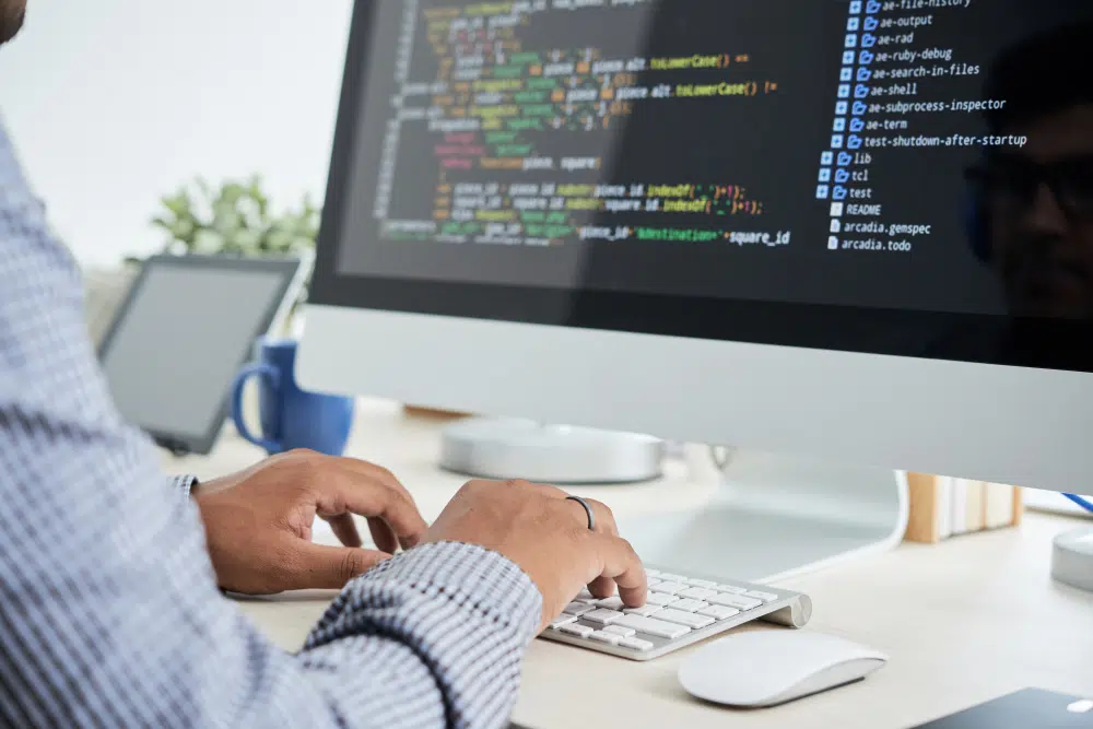 A person types on a white keyboard at a modern workstation with a large monitor displaying lines of code and a file directory. The desk includes a tablet, a blue coffee mug, and a small plant, creating a clean and organised software‑development workspace.