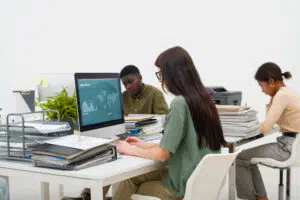 Three people work at desks in a modern office filled with stacks of documents and folders. The person in the foreground is viewing a computer screen displaying “ANNUAL STATISTICS” with a world map and a line chart labeled “NEW USERS.” The scene highlights data analysis and a busy administrative work environment.
