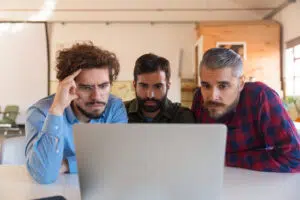 Three people sit closely together at a table in a warm, wood‑toned office space, looking intently at a laptop screen. The person on the left wears glasses and a blue shirt, the person in the middle has a beard and a dark shirt, and the person on the right has grey hair, a beard, and a red‑and‑blue plaid shirt. They appear focused and engaged in collaborative work.