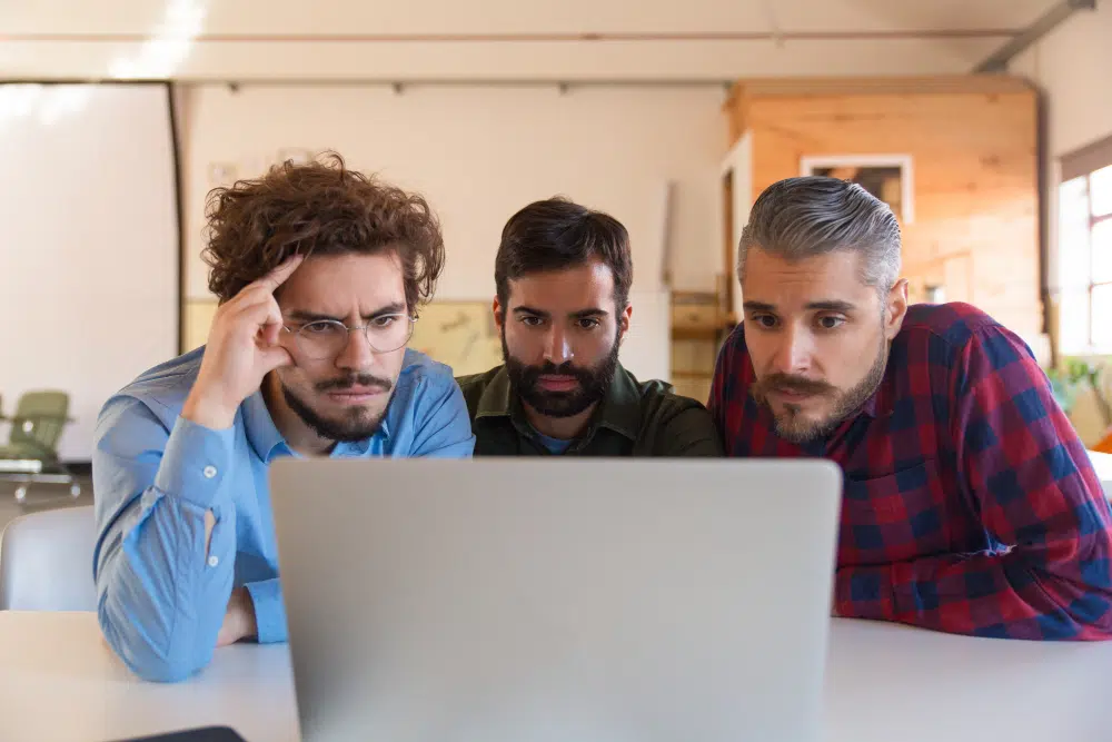 Three people sit closely together at a table in a warm, wood‑toned office space, looking intently at a laptop screen. The person on the left wears glasses and a blue shirt, the person in the middle has a beard and a dark shirt, and the person on the right has grey hair, a beard, and a red‑and‑blue plaid shirt. They appear focused and engaged in collaborative work.