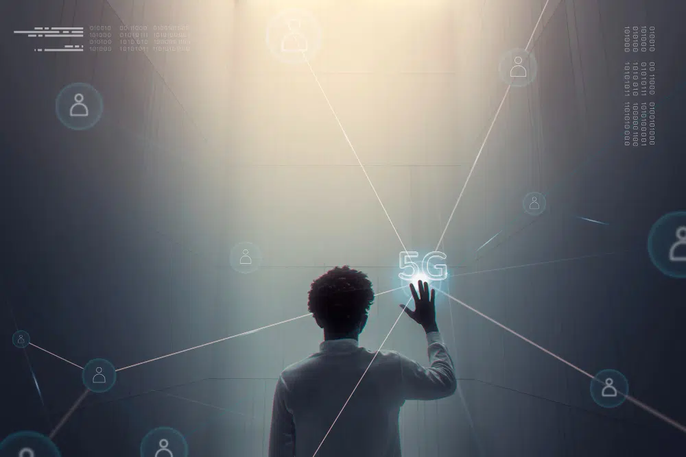 A person stands in a dark, futuristic room reaching toward a glowing “5G” projection. Network icons and connecting lines float around them, with binary code on the walls, creating a visual representation of advanced digital connectivity.