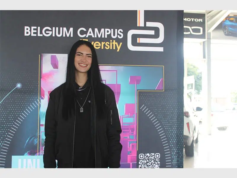A person stands in front of a colourful Belgium Campus iTversity promotional backdrop featuring geometric shapes in blue, pink, and purple, along with the institution’s name in white and yellow text. A QR code appears near the bottom right of the display. Cars are visible in the background, suggesting the photo was taken at an indoor event or exhibition.