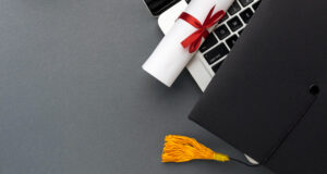 A rolled-up diploma tied with a red ribbon rests on a laptop keyboard, with a black graduation cap and yellow tassel placed beside it on a smooth grey surface, symbolising academic achievement and digital learning.