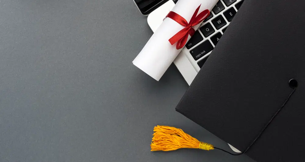 A rolled-up diploma tied with a red ribbon rests on a laptop keyboard, with a black graduation cap and yellow tassel placed beside it on a smooth grey surface, symbolising academic achievement and digital learning.