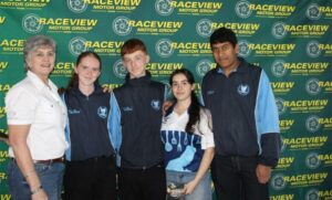 Five people posing in front of a Raceview Motor Group backdrop; an older woman on the left with four young team members in blue jackets beside her.