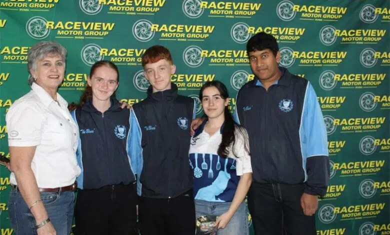 Five people posing in front of a Raceview Motor Group backdrop; an older woman on the left with four young team members in blue jackets beside her.