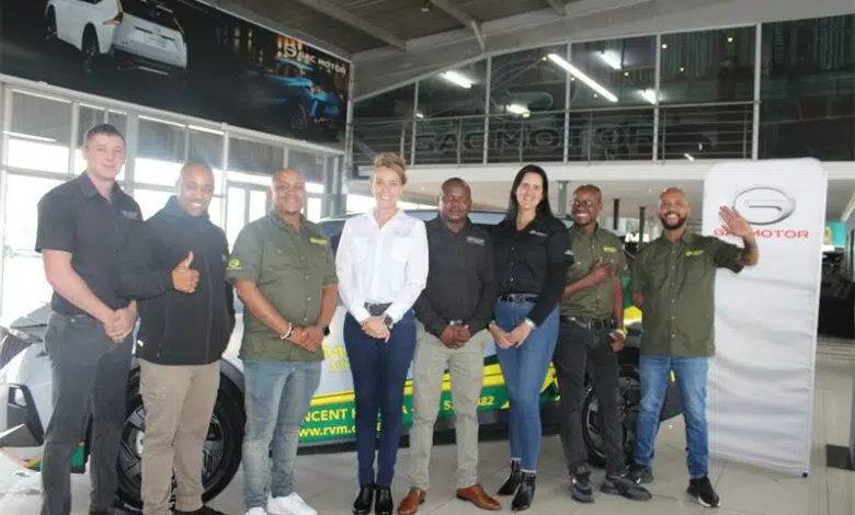 A group of seven people stand in a modern GAC Motor showroom, posing in front of a branded vehicle with green and yellow graphics. Large glass windows, high ceilings, and car posters frame the space, giving the scene a professional, automotive‑industry atmosphere.