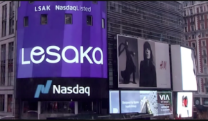 A vibrant Times Square scene featuring tall digital billboards, including a large curved purple display reading “Lesaka” with “LSAK NasdaqListed” above it, positioned above the Nasdaq logo. To the right is an H&M advertisement showing two black‑and‑white fashion portraits. Additional smaller screens display colourful ads and city visuals against the busy urban backdrop.