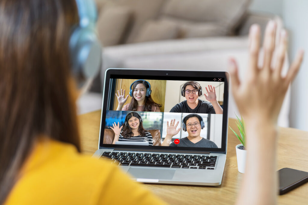 A person wearing headphones sits at a desk, raising a hand in greeting during a video call on a laptop. Four people appear on the laptop screen in a grid, each wearing headphones and waving back. A small potted plant and a smartphone sit beside the laptop, creating a casual remote‑meeting setup.