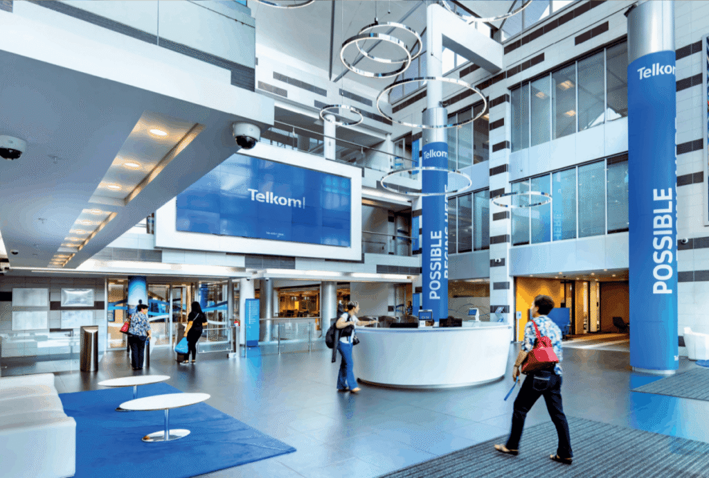 A spacious, modern Telkom corporate lobby with high ceilings, glass walls, and circular ring lights suspended from above. A white circular reception desk sits in the centre, with several people walking or standing nearby. Blue pillars display the word “POSSIBLE” vertically, and a large digital screen shows the Telkom logo on a bright blue background. The space feels open, professional, and contemporary.