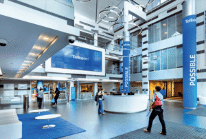 A spacious, modern Telkom corporate lobby with high ceilings, glass walls, and circular ring lights suspended from above. A white circular reception desk sits in the centre, with several people walking or standing nearby. Blue pillars display the word “POSSIBLE” vertically, and a large digital screen shows the Telkom logo on a bright blue background. The space feels open, professional, and contemporary.
