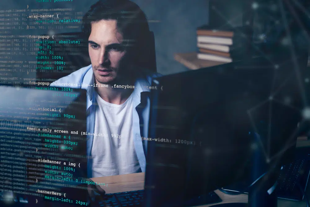 A person sits at a desk working on a computer with multiple monitors, while lines of CSS code are overlaid in the foreground, creating a layered visual of focused programming and digital development.