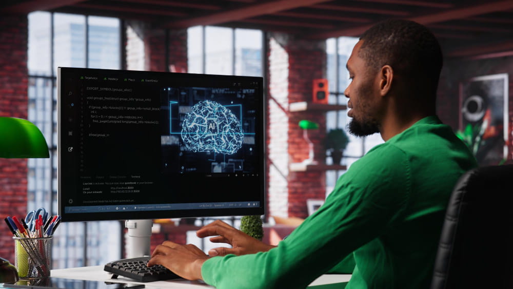 A person sits at a desk in a modern office, working on a computer displaying programming code on one side of the screen and a glowing digital brain labelled “AI” on the other. The workspace includes a keyboard, mouse, green desk lamp, and a container of pens, with large windows and shelves in the background creating a contemporary tech‑focused setting.