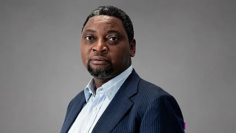 A person wearing a dark blue pinstripe suit jacket and a light blue collared shirt poses against a plain grey background in a formal portrait.