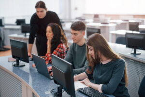 A classroom-style computer lab with several students working at desktop computers. Three people sit at a curved desk using laptops and monitors, while another person stands nearby offering assistance. Rows of computer stations fill the bright, modern room, creating a focused learning environment.