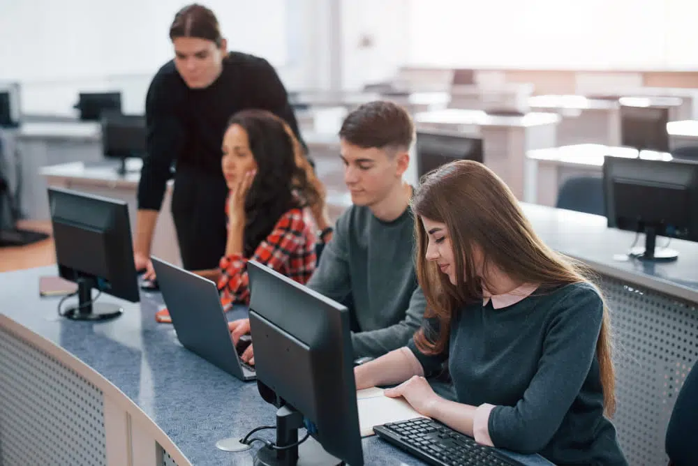 A classroom-style computer lab with several students working at desktop computers. Three people sit at a curved desk using laptops and monitors, while another person stands nearby offering assistance. Rows of computer stations fill the bright, modern room, creating a focused learning environment.