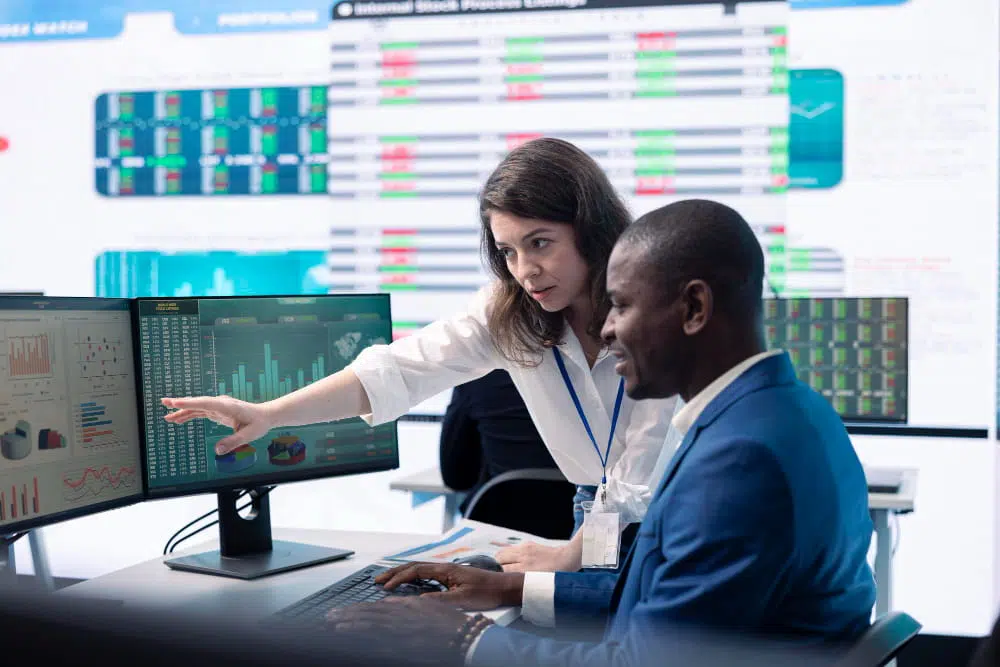 Two people sit together at a desk in a modern office, analysing data on multiple computer monitors. The screens display colourful bar graphs, pie charts, line graphs, and numerical tables. One person points at a chart while the other types on a keyboard. Large digital displays in the background show additional financial or analytical data, creating a high‑tech, collaborative workspace atmosphere.