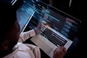 A person sits at a desk working on a laptop displaying lines of HTML and JavaScript code, including React script references. The scene focuses on the screen and hands typing, highlighting software development work.