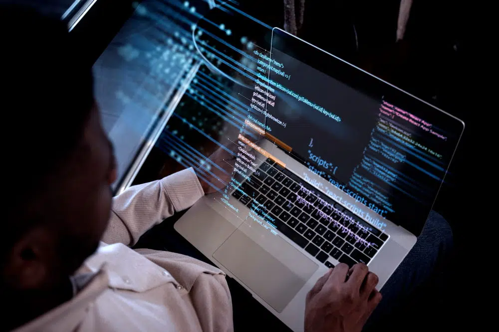 A person sits at a desk working on a laptop displaying lines of HTML and JavaScript code, including React script references. The scene focuses on the screen and hands typing, highlighting software development work.