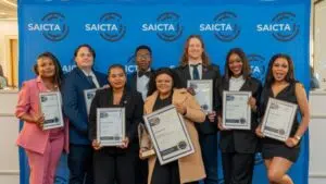A group of eight people stand in front of a blue SAICTA-branded backdrop, each holding a framed certificate. One person in the front centre also holds a trophy. They are dressed in smart or formal attire and are posing together for a celebratory photo, suggesting they have received awards or recognition.