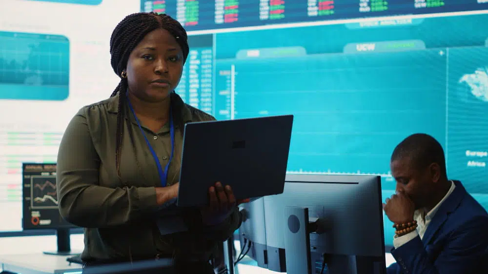 Woman standing with a laptop in a modern control room, man seated at a desk looking at a monitor in the background.