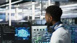 A person in a white lab coat stands in a high‑tech workspace, facing multiple computer monitors. The screens display complex digital graphics, including a glowing blue brain diagram and a network flowchart. The individual has headphones around their neck, suggesting technical or research work in an advanced computing or AI environment.